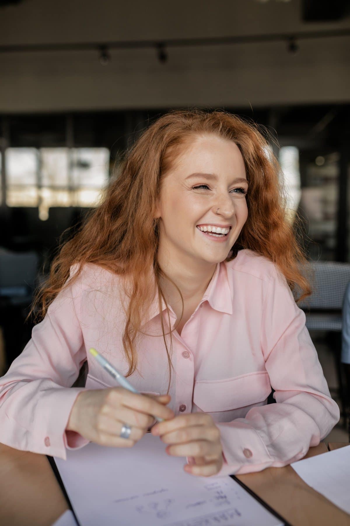 Rapariga ruiva de camisa rosa a rir, à mesa de trabalho